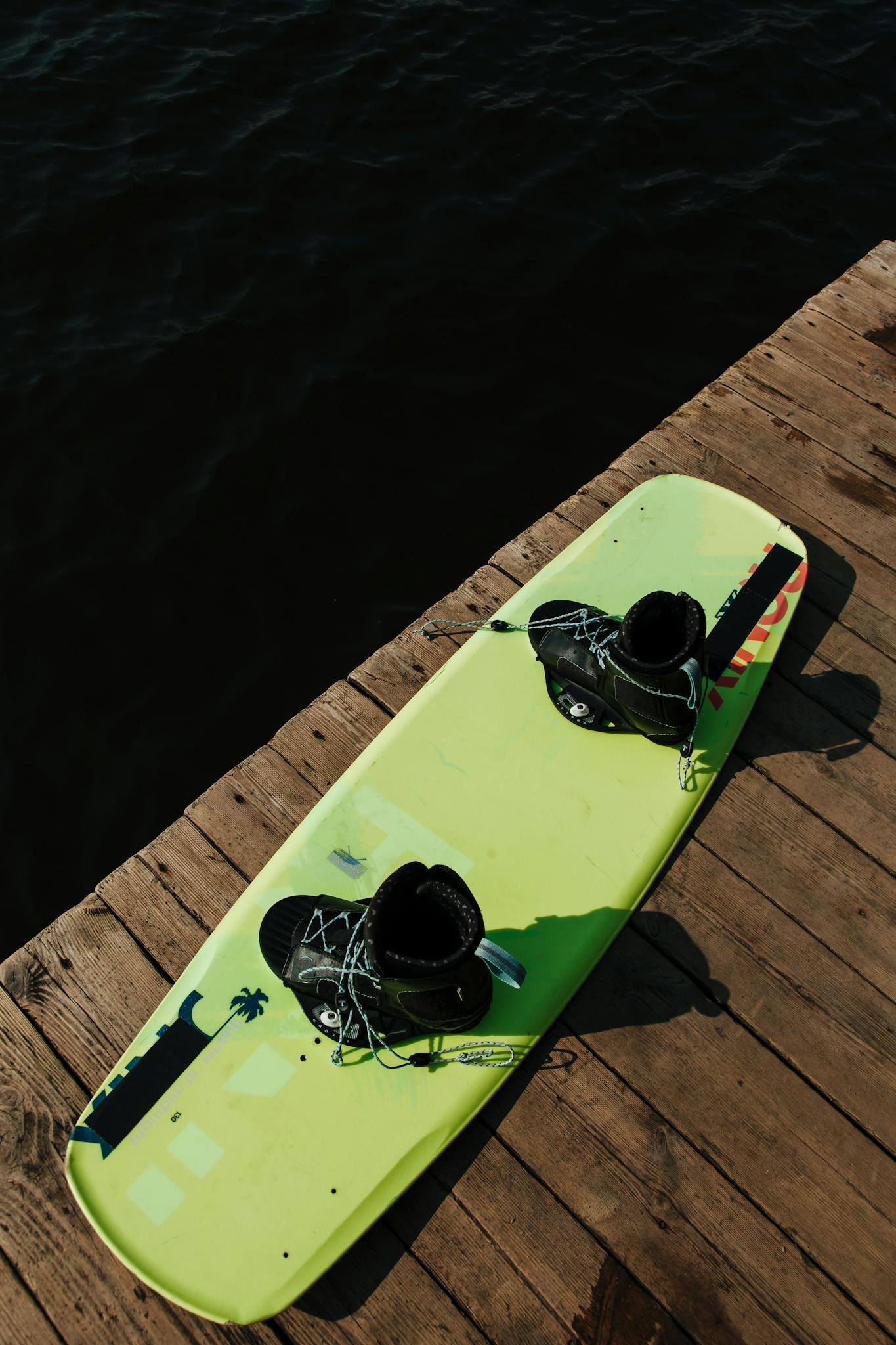 A vibrant wakeboard with bindings on a wooden jetty by calm water, ready for sports action.